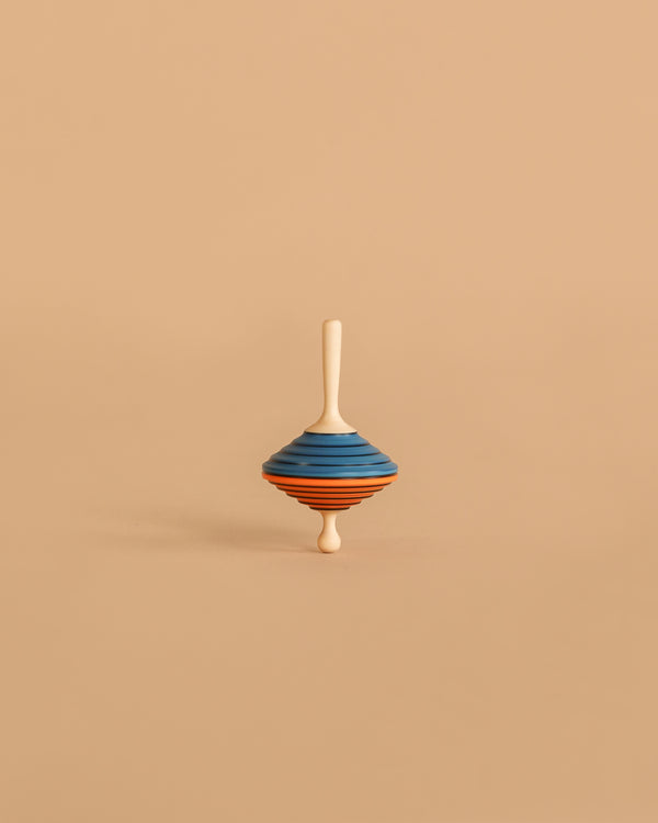 The Mader Tukanino Spinning Top - Blue features a maple wood top with blue and orange stripes, standing upright on a beige background and recalling the charm of classic Austrian toys.