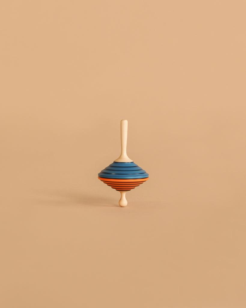 The Mader Tukanino Spinning Top - Blue features a maple wood top with blue and orange stripes, standing upright on a beige background and recalling the charm of classic Austrian toys.