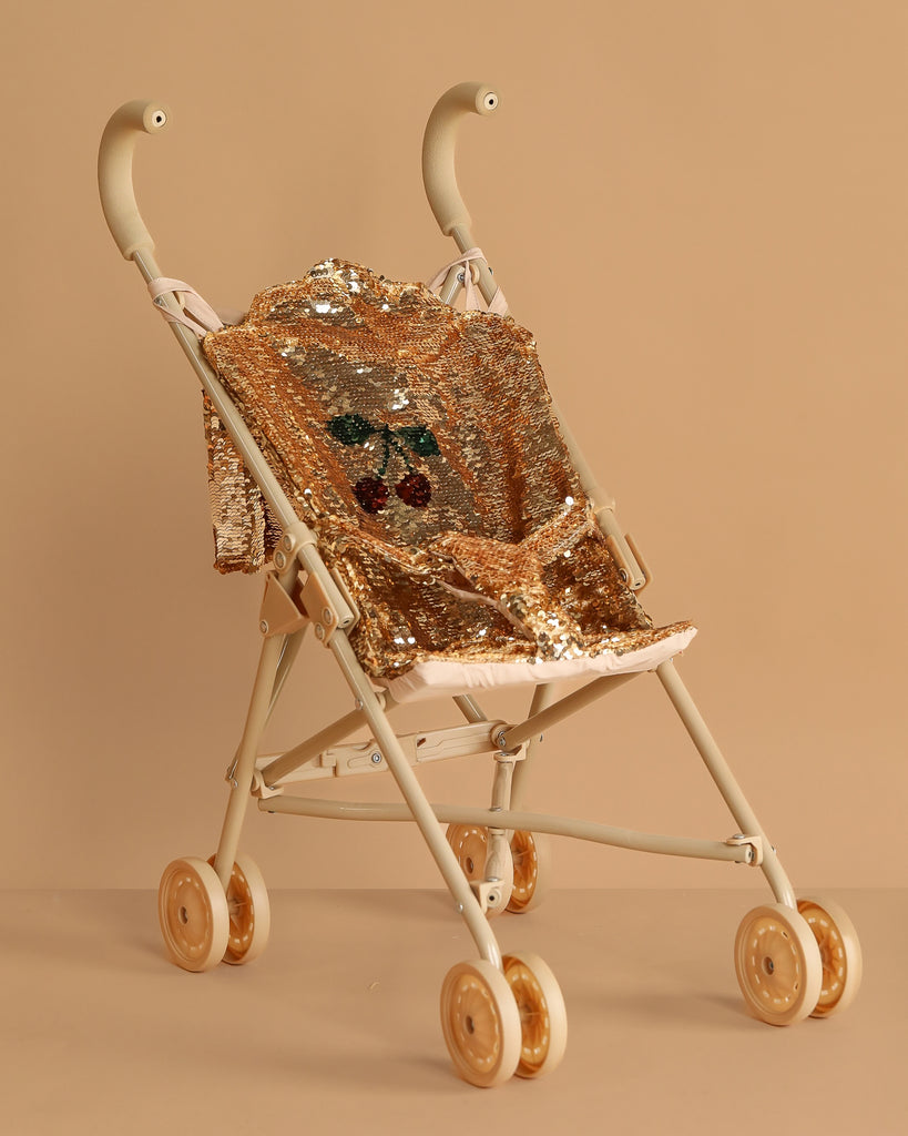 The Konges Sloejd Sparkly Sequin Doll Stroller - Golden Cherry features gold wheels and shiny gold sequin fabric with a cherry design on a matching beige background.