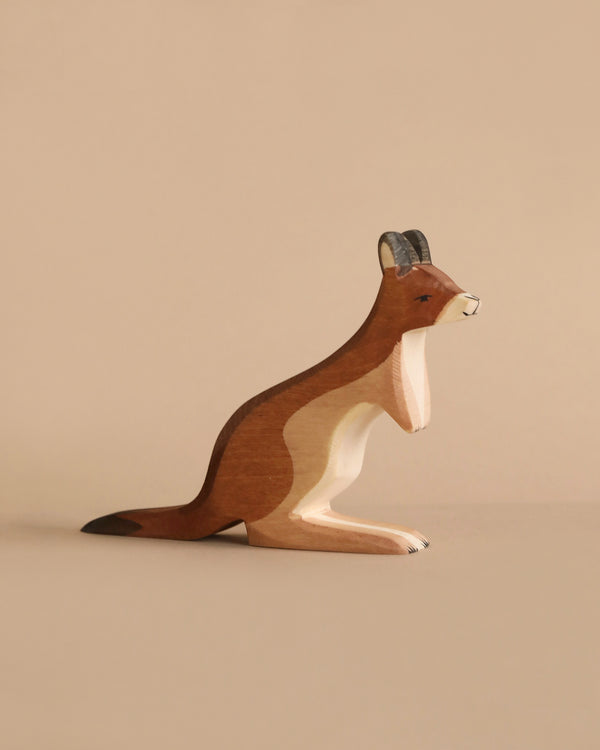 A handcrafted wooden figurine of a kangaroo, featuring detailed carving and a natural wood grain finish, stands against a plain, light brown background. Made from sustainable materials, the Ostheimer Kangaroo Father is depicted in a relaxed position with its tail extended behind it.