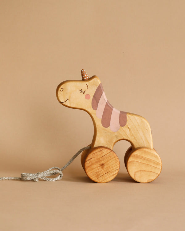 Handmade Wooden Unicorn Pull Toy made from sustainably harvested birch wood, featuring painted details such as eyes and a striped saddle using natural milk paint, with wheels and a pull string on a beige background.