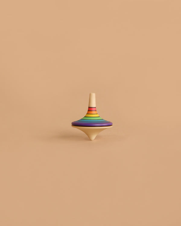 The Mader Rallye Rainbow Spinning Top, Small - Purple, featuring vibrant rainbow stripes and crafted from smooth beech wood, rests on a simple beige background.
