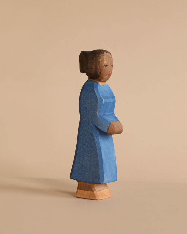 A wooden figurine of a woman stands against a beige background. The figure is carved in a simple, minimalist style and painted wearing a long blue dress. Featuring a modest hairstyle with hair pulled back and a calm expression, this piece embodies the essence of Ostheimer Mother, Blue Dress for imaginative play.
