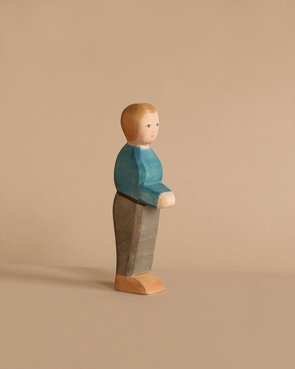 A handcrafted wooden figurine of a person stands against a beige background. Made by Ostheimer in Germany, the Ostheimer Father, Blue Sweater is painted with a blue top, gray pants, and beige shoes. Its plain, simple design conveys a minimalist aesthetic. The figurine has a neutral expression and is looking slightly forward.