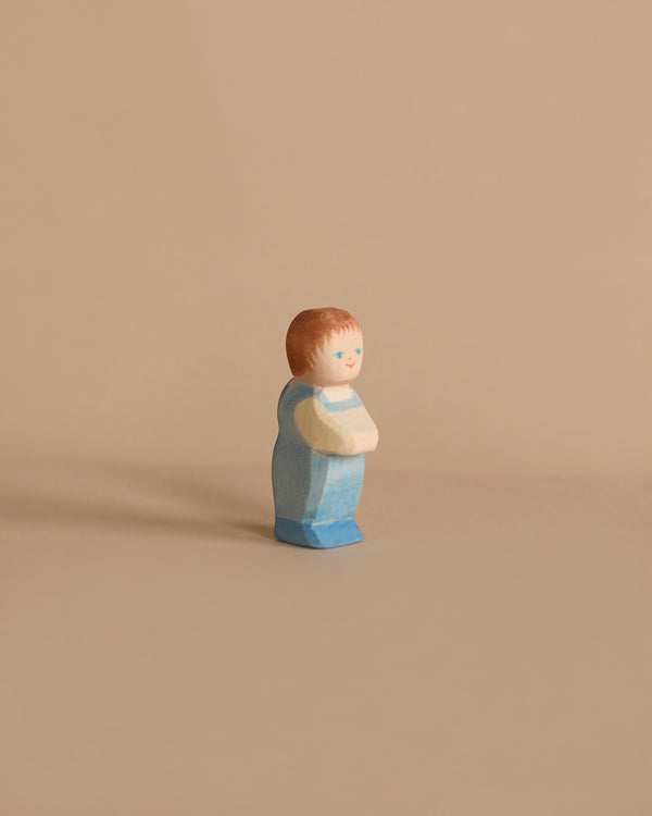 A small, hand-painted Ostheimer Toddler figurine of a person with light brown hair, wearing a blue outfit, stands against a beige background. Made from sustainably sourced materials, the figure’s hands are clasped in front and it has a serene facial expression with simple, minimalist features.