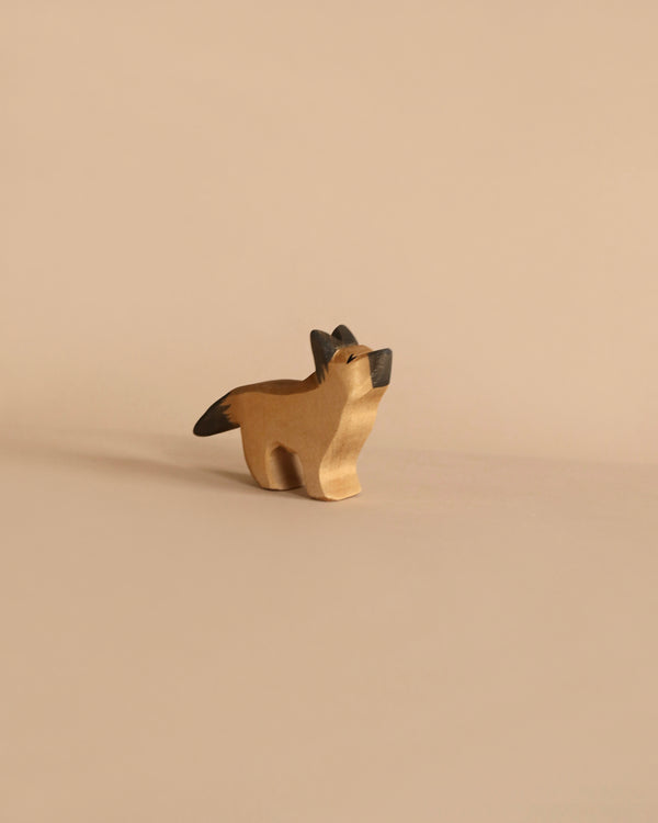 A small, handcrafted Ostheimer German Shepherd, Small with a minimalist design stands against a beige background. Reminiscent of Ostheimer wooden toys, it has a smooth, light brown body with black accents on its ears and tail. The German Shepherd is positioned facing slightly upward, giving it a playful appearance.