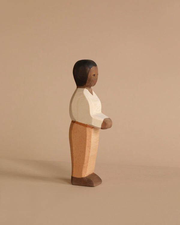 A wooden figurine of a person with short black hair, wearing a white shirt and tan pants, stands against a beige background. The figurine has minimal facial features and a simple, handcrafted appearance, reminiscent of the Ostheimer Father, Cream Sweater toy, perfect for imaginative play.