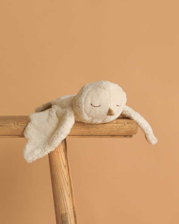 The Senger Naturwelt Cuddly Animal - Owl, a soft cream-colored stuffed owl with closed eyes, is draped over a wooden ladder set against a plain light brown background.