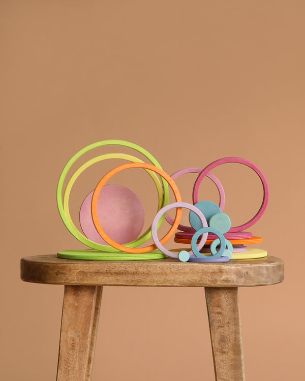 Grimm's Concentric Circles and Rings Building Set - Pastel features colorful wooden rings and discs of varying sizes, artfully stacked on a stool against a light brown backdrop, creating a playful and balanced display.