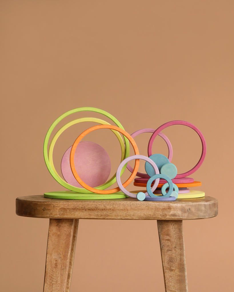 Grimm's Concentric Circles and Rings Building Set - Pastel features colorful wooden rings and discs of varying sizes, artfully stacked on a stool against a light brown backdrop, creating a playful and balanced display.