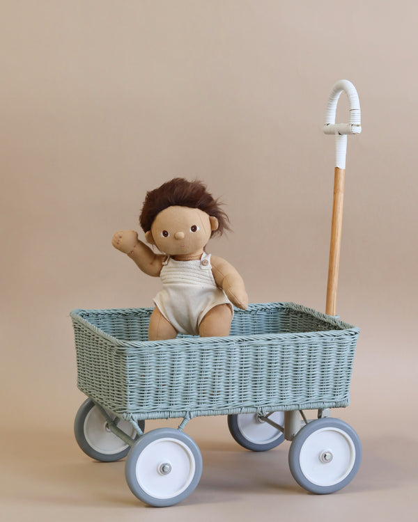 A plush doll with brown hair sits in a blue Olli Ella Rattan Wonder Wagon - Vintage Blue with white plastic wheels, against a neutral beige background. The wagon handle is wooden and white.