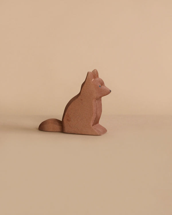A small, carved wooden figurine of a fox sits against a beige background. The fox is in a sitting position, facing to the right, with a simple, minimalist design and smooth finish. Made from sustainably sourced materials, this piece embodies the quality of handcrafted wooden toys by Ostheimer Shepherd's Dog.