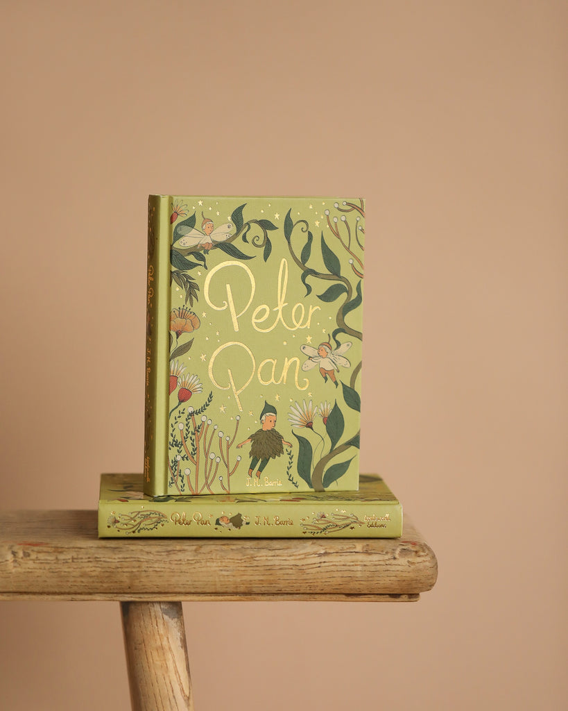 Two copies of the Peter Pan Collector's Edition by J.M. Barrie sit on a wooden stool. The whimsical covers feature green vines and tiny fairies on a light green background, with the title elegantly displayed in gold lettering against a soft beige backdrop.