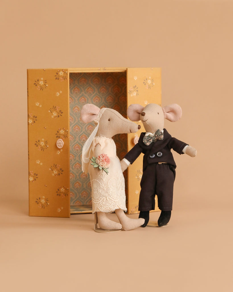 The Maileg Wedding Mice Couple in Box, Mum and Dad, features two fabric mice dressed as bride and groom with lace and veil, and suit with bow tie, posed before a floral-patterned box against a beige background.