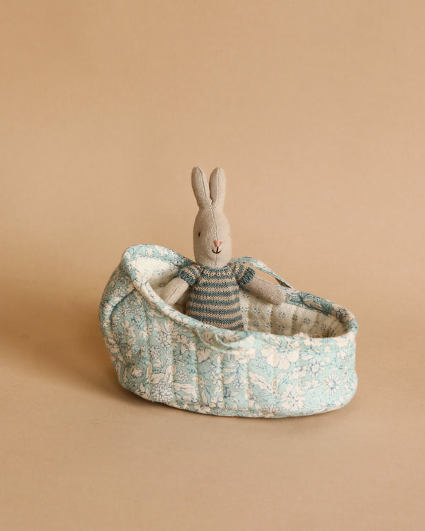 A Maileg Rabbit in Carry Cot, Micro - Blue with long ears sitting in a fabric-covered oval carry cot, against a plain beige background.
