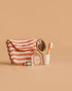 A striped pouch, a small perfume bottle, a tube of lipstick, a gold handheld mirror, and a pink toothbrush from the Maileg Powder Room Set in a white cup are arranged on a beige background.