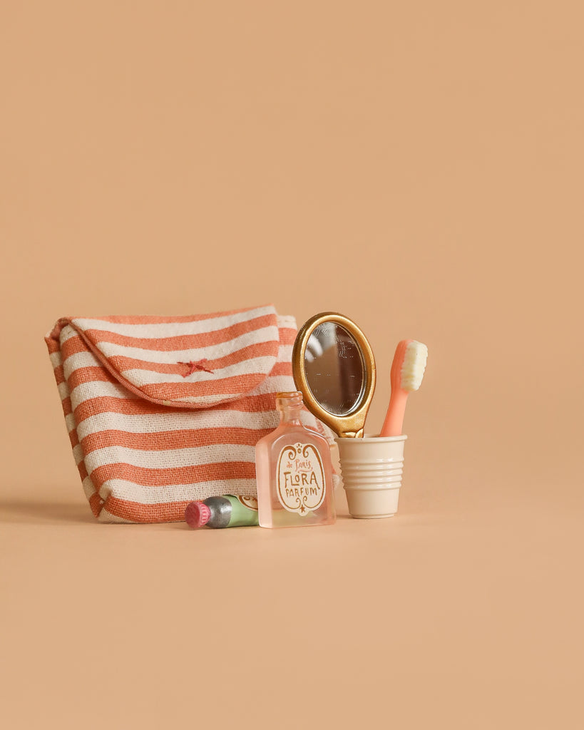 A striped pouch, a small perfume bottle, a tube of lipstick, a gold handheld mirror, and a pink toothbrush from the Maileg Powder Room Set in a white cup are arranged on a beige background.
