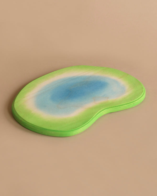 A stack of kidney-shaped coasters with a gradient design resembling a watercolor painting, featuring a green outer edge blending into a blue center. These Ostheimer Ponds are set against a simple beige background.