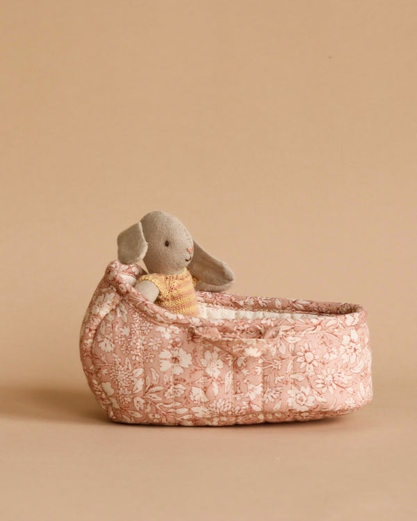 A small plush Maileg Rabbit in Carry Cot, Micro - Rose, with long ears, sits in a pink, floral-patterned fabric basket. The background is a plain beige color, creating an inviting playset for children.