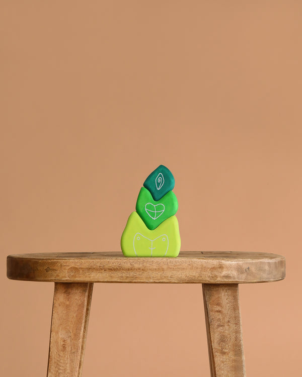 Three green Grimm's Tower Leaves Building Blocks—handpainted lime wood stones with white leaf symbols—are stacked on a wooden stool against a plain beige background.
