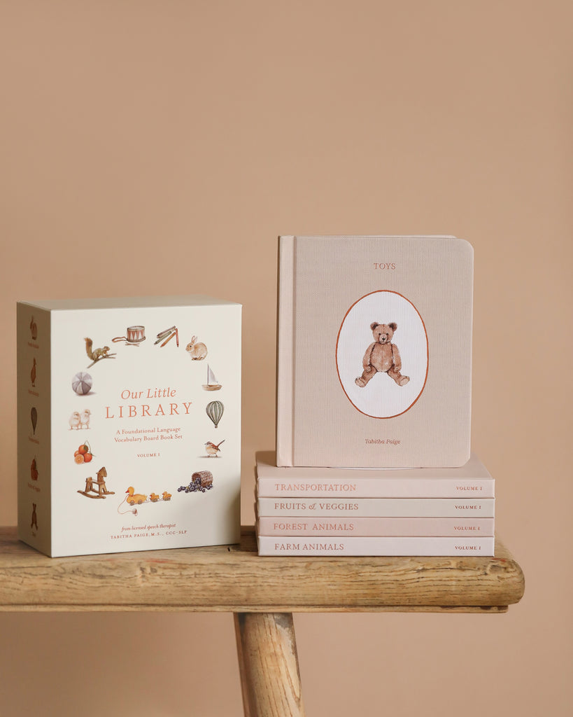 A set of baby board books from the Our Little Library Book Box Set rests on a wooden bench against a beige background. Perfect for speech therapists, it includes themed titles like 'First Words,' 'Transportation,' 'Fruits & Veggies,' 'Forest Animals,' and 'Farm Animals.'.