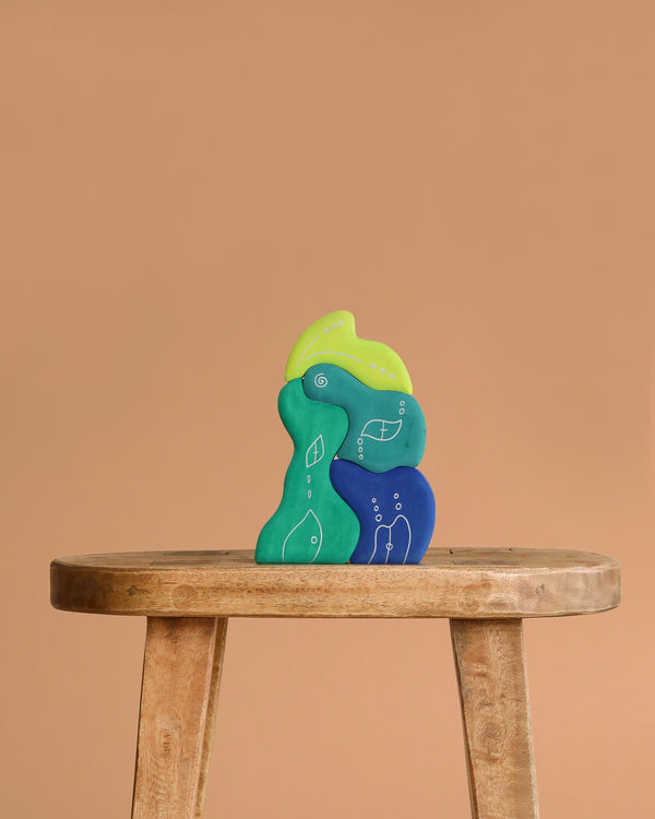 Grimm's Casa Aqua Building Blocks—abstract, colorful pieces in green, blue, and teal with white line drawings—are arranged on a wooden stool against a plain beige background. Their irregular shapes inspire imaginative play.