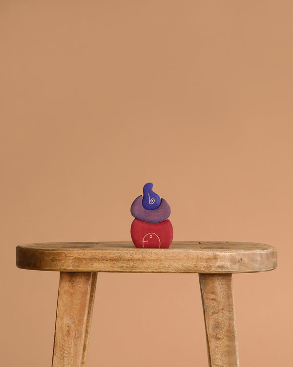 A trio of Grimm's Small Bakery Building Blocks—blue, purple, and red—are stacked atop a rustic wooden stool, standing out against a plain beige background.