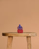 A trio of Grimm's Small Bakery Building Blocks—blue, purple, and red—are stacked atop a rustic wooden stool, standing out against a plain beige background.