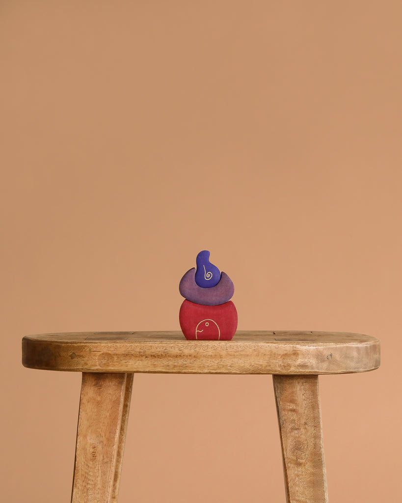 A trio of Grimm's Small Bakery Building Blocks—blue, purple, and red—are stacked atop a rustic wooden stool, standing out against a plain beige background.