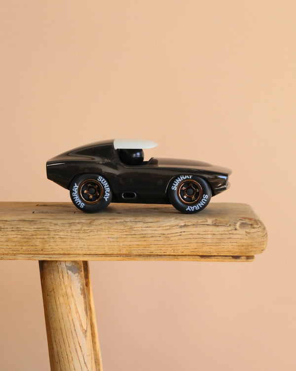 The Playforever Leadbelly Skeeter Car in Black, featuring white tire lettering, rests on a wooden surface with a plain beige background.