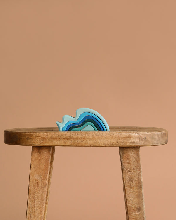 The Grimm's Ocean Waves, a small blue abstract wave-shaped toy, rests on a simple wooden stool against a plain beige background.