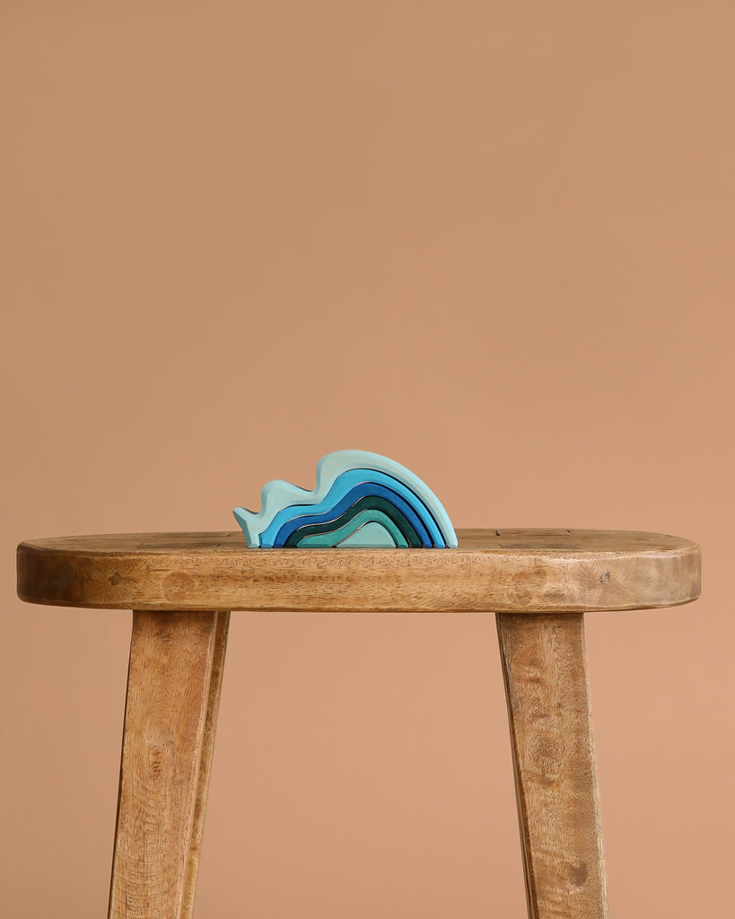 The Grimm's Ocean Waves, a small blue abstract wave-shaped toy, rests on a simple wooden stool against a plain beige background.