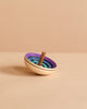 The Mader UFO Flora Spinning Top - Purple, made of maple wood and featuring ombre blue and purple rings with a brown handle, rests on a light beige surface against a matching background.