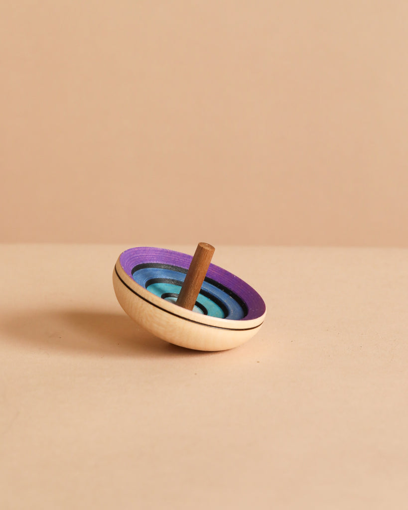 The Mader UFO Flora Spinning Top - Purple, made of maple wood and featuring ombre blue and purple rings with a brown handle, rests on a light beige surface against a matching background.