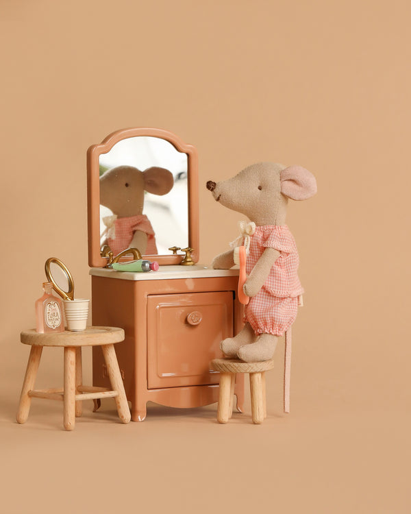 The Maileg Bathroom Set features a Little Sister Mouse in a pink gingham outfit seated at a peach-colored vanity with mirror, while a wooden stool nearby displays accessories like a clock, cup, and hand mirror against a light brown backdrop.