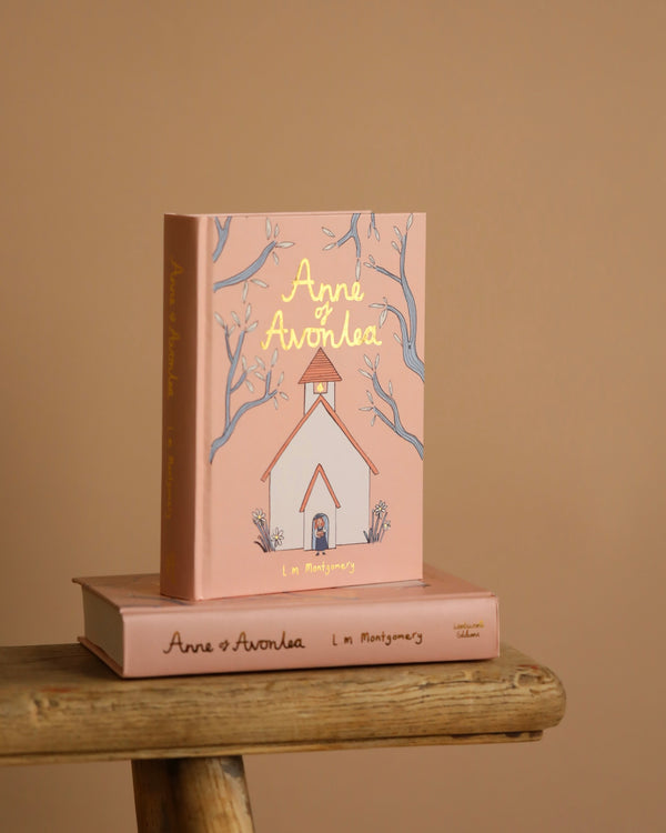 Two "Anne of Avonlea | Collector's Edition | Hardcover" books are shown. One features a pastel pink cover with a schoolhouse and tree illustration, depicting Anne as a teacher, standing upright. The other is lying flat on a wooden surface.