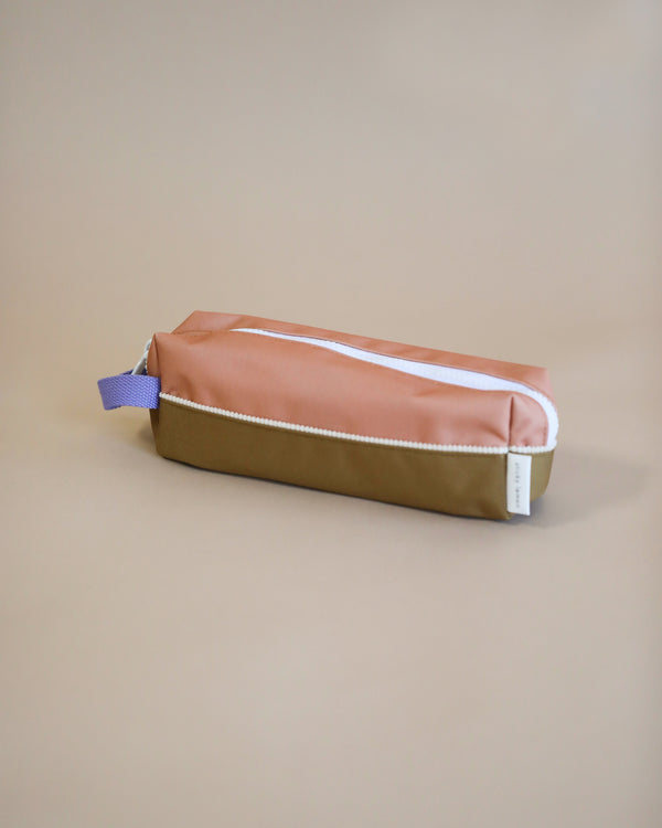 A Sticky Lemon pencil case, with the top half in Harvest Moon and the bottom in Soil Green, resting on a beige surface. It features a YKK zipper and a blue loop tab on.