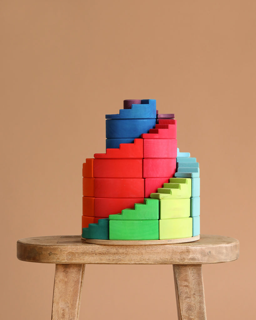 Grimm's Counterrotating Stepped Spiral, a set of colorful, semi-circular wooden blocks, is stacked in a spiral on a round stool against a beige background, inviting creative play and supporting fine motor skill development.