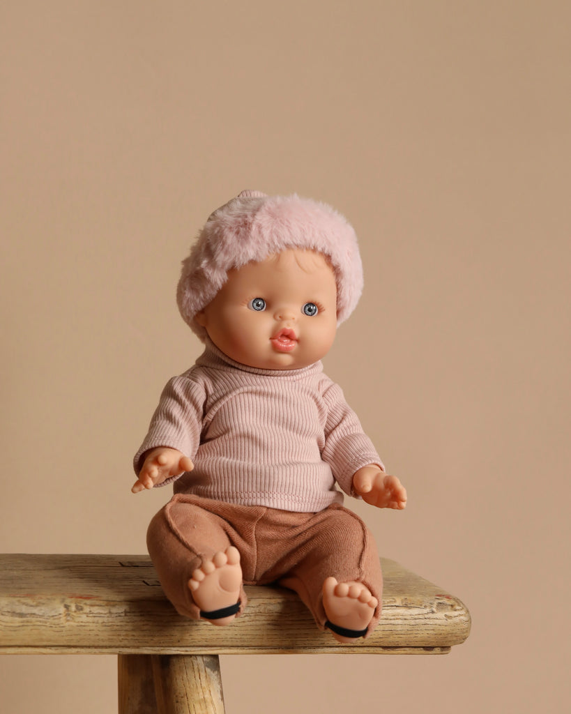 A Minikane Vintage Baby Doll (13") - Lily with pink hat and light brown clothes sits on a wooden stool against a beige background, showcasing blue eyes and a neutral expression while emitting a subtle natural vanilla scent.