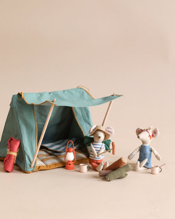 Two toy mice dressed in cozy clothes, sitting beside a Maileg Happy Camper Tent with a sleeping bag, blanket, and a lantern, on a neutral background.