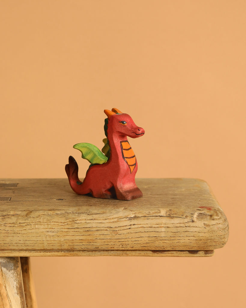 The Bumbu Handmade Wooden Baby Dragon is a small red figurine with green wings and orange details, resting on a rustic wooden surface—ideal for fantasy play—set against a plain peach background.