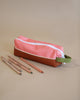 A Sticky Lemon Pencil Case in Flower Pink and Willow Brown, crafted from recycled PET bottles and equipped with a sturdy green strap, rests on a light brown surface. Nearby, four colored pencils are casually arranged. The plain background subtly highlights the pencil case and pencils.