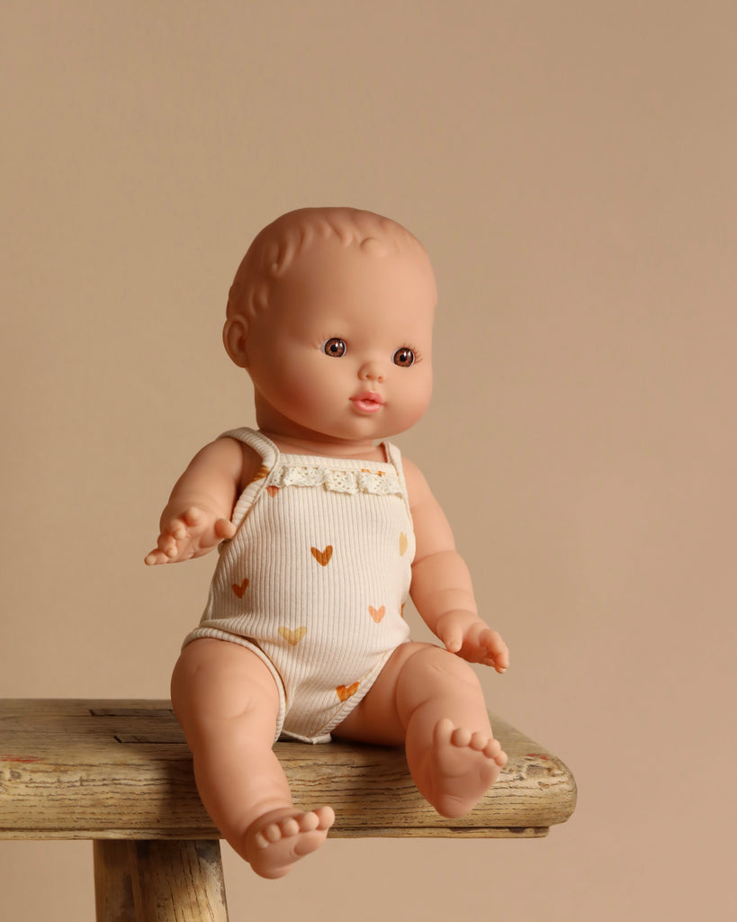 The Minikane Vintage Baby Doll (13") - Faustine with Clothing sits on a wooden stool in a white romper with brown hearts. Crafted from phthalate-free vinyl, it's perfect for imaginative play against a warm beige backdrop.