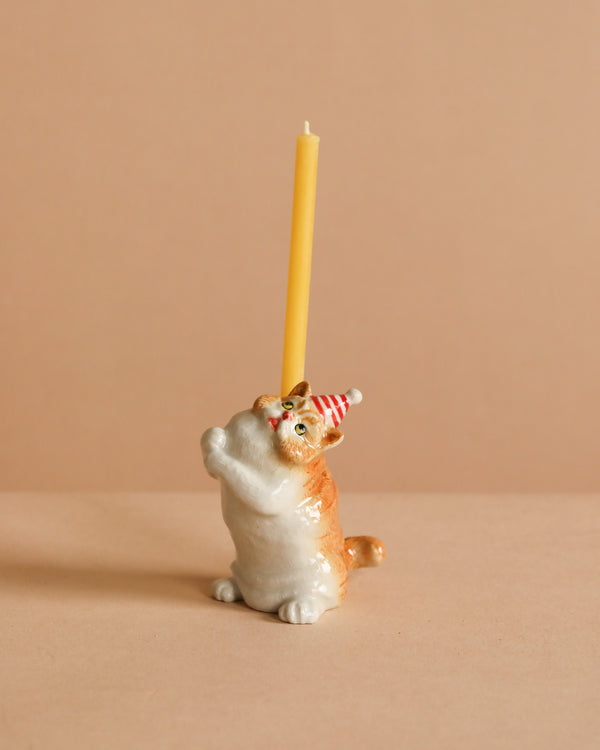 The Exotic Shorthair Cat Cake Topper features a ceramic cat figurine in a red and white party hat, holding a yellow candle upright against a beige background—perfect for adding whimsy to any cat-themed celebration.