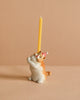 The Exotic Shorthair Cat Cake Topper features a ceramic cat figurine in a red and white party hat, holding a yellow candle upright against a beige background—perfect for adding whimsy to any cat-themed celebration.