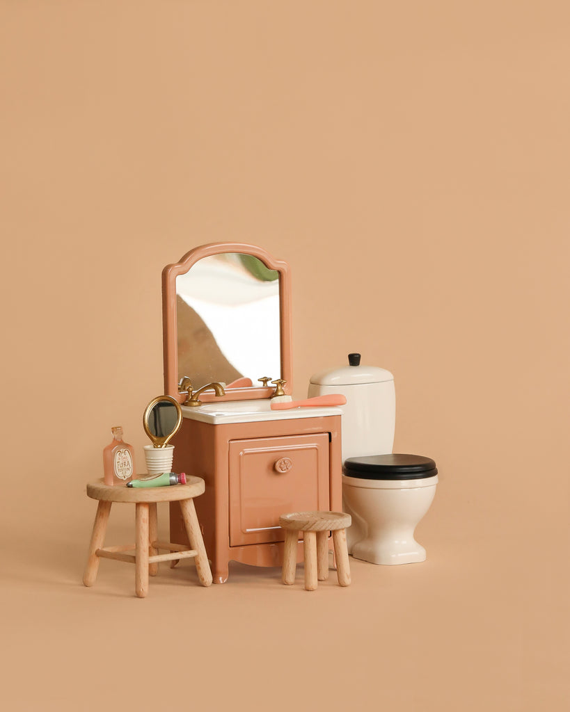 A miniature bathroom scene featuring the Maileg Bathroom Set, including a white toilet with black lid and two wooden stools. The toothbrush set and soap rest on a stool, all displayed against a solid beige background.