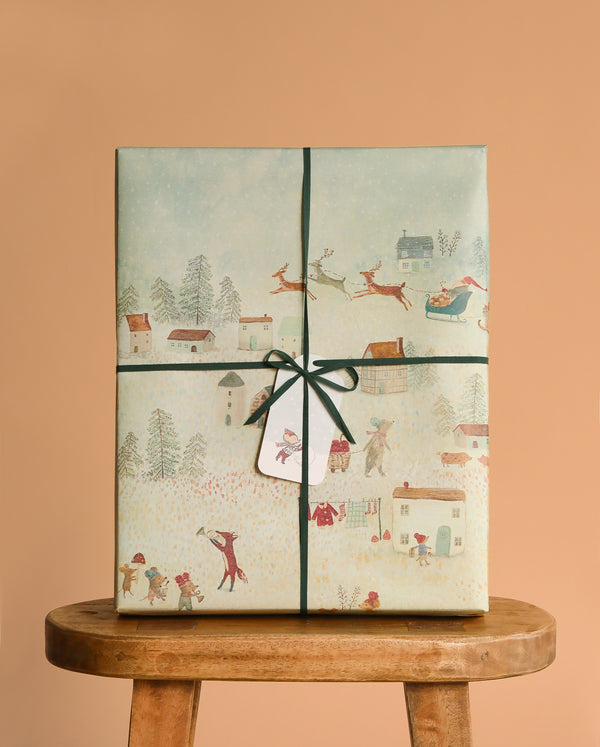 The Maileg Mystery Box - Christmas, wrapped in festive paper with winter village scenes and Santa with reindeer, tied with green string and a gift tag, brings Maileg Christmas charm on a wooden stool against a beige background.