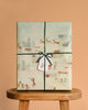 The Maileg Mystery Box - Christmas, wrapped in festive paper with winter village scenes and Santa with reindeer, tied with green string and a gift tag, brings Maileg Christmas charm on a wooden stool against a beige background.