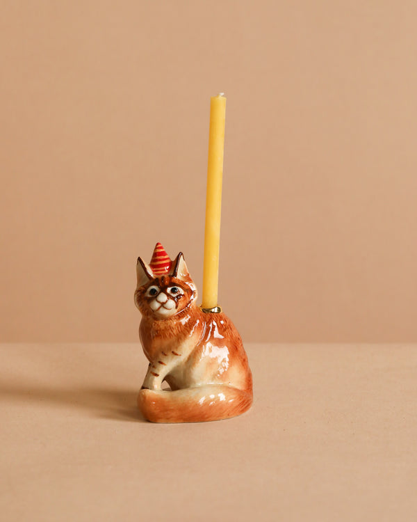 The Maine Coon Cat Cake Topper is a lifelike ceramic figurine with a party hat, holding a yellow candle on its back—ideal for cat-themed parties, shown against a plain beige background.