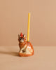 The Maine Coon Cat Cake Topper is a lifelike ceramic figurine with a party hat, holding a yellow candle on its back—ideal for cat-themed parties, shown against a plain beige background.
